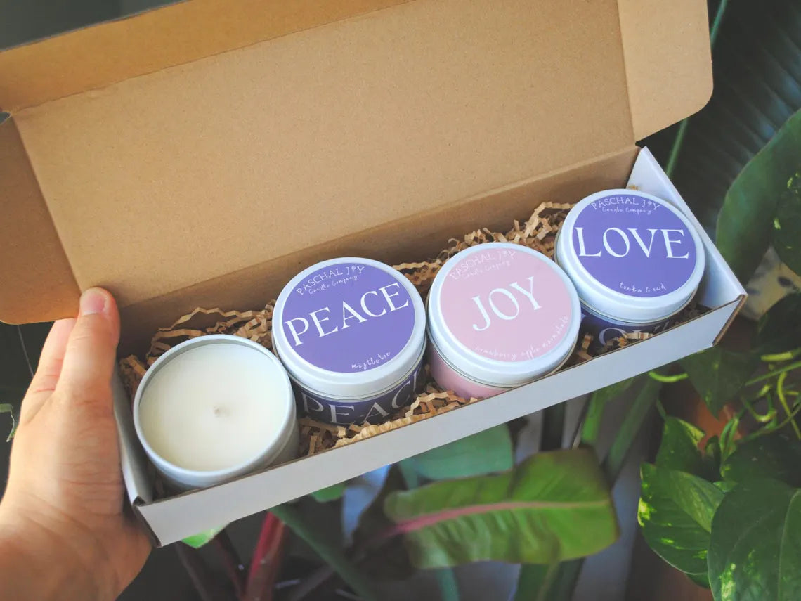 Four small candles with text on lids in a box, held by a hand with plants in the background.
