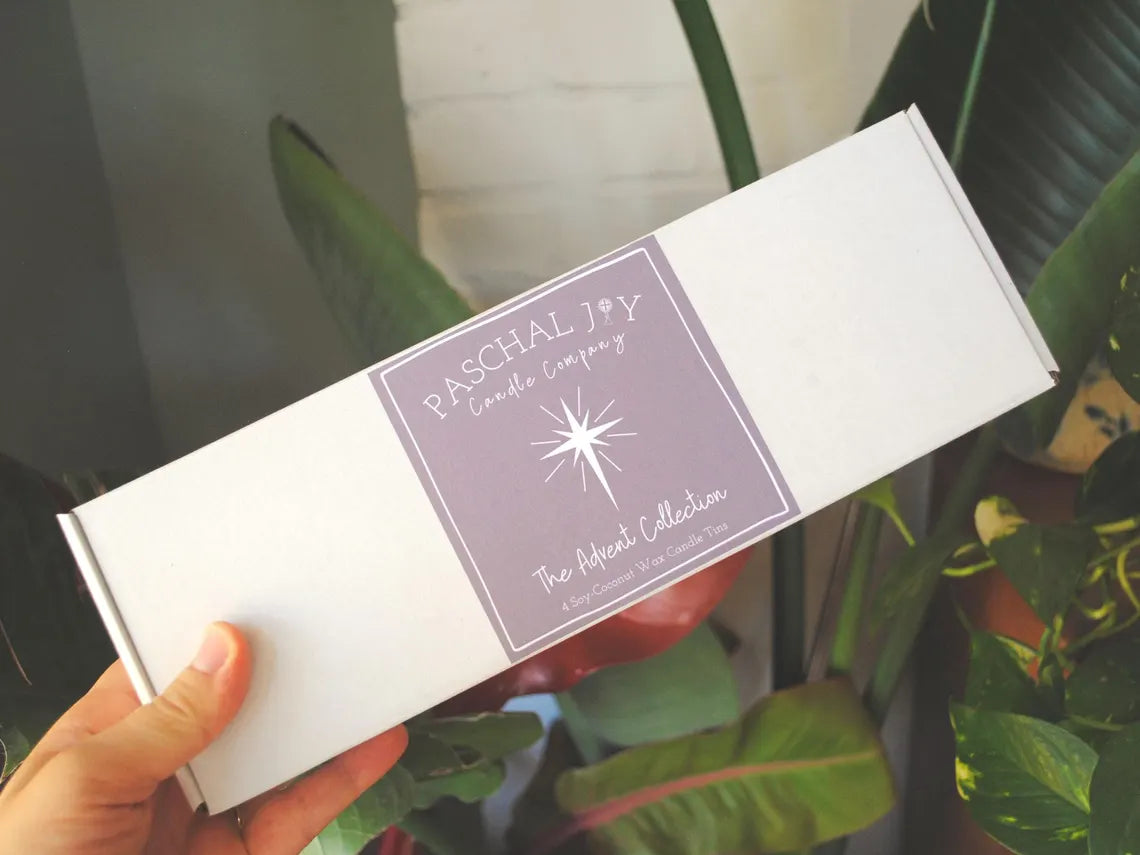 Hand holding a box with 'Paschal Joy Candle Company' label against a plant background