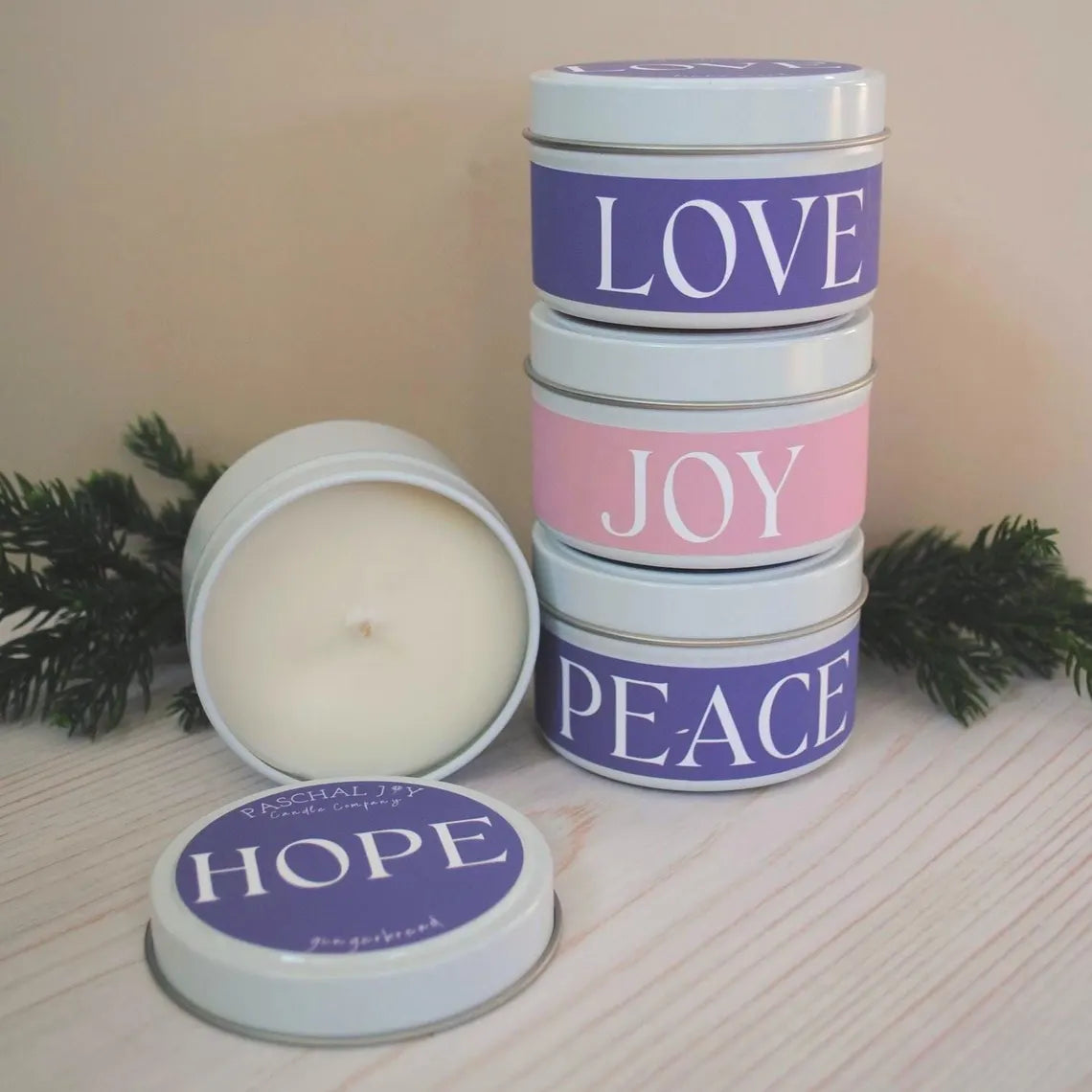 Stack of small candles in metal tins labeled 'Love', 'Joy', and 'Peace' on a wooden surface with a soft background.