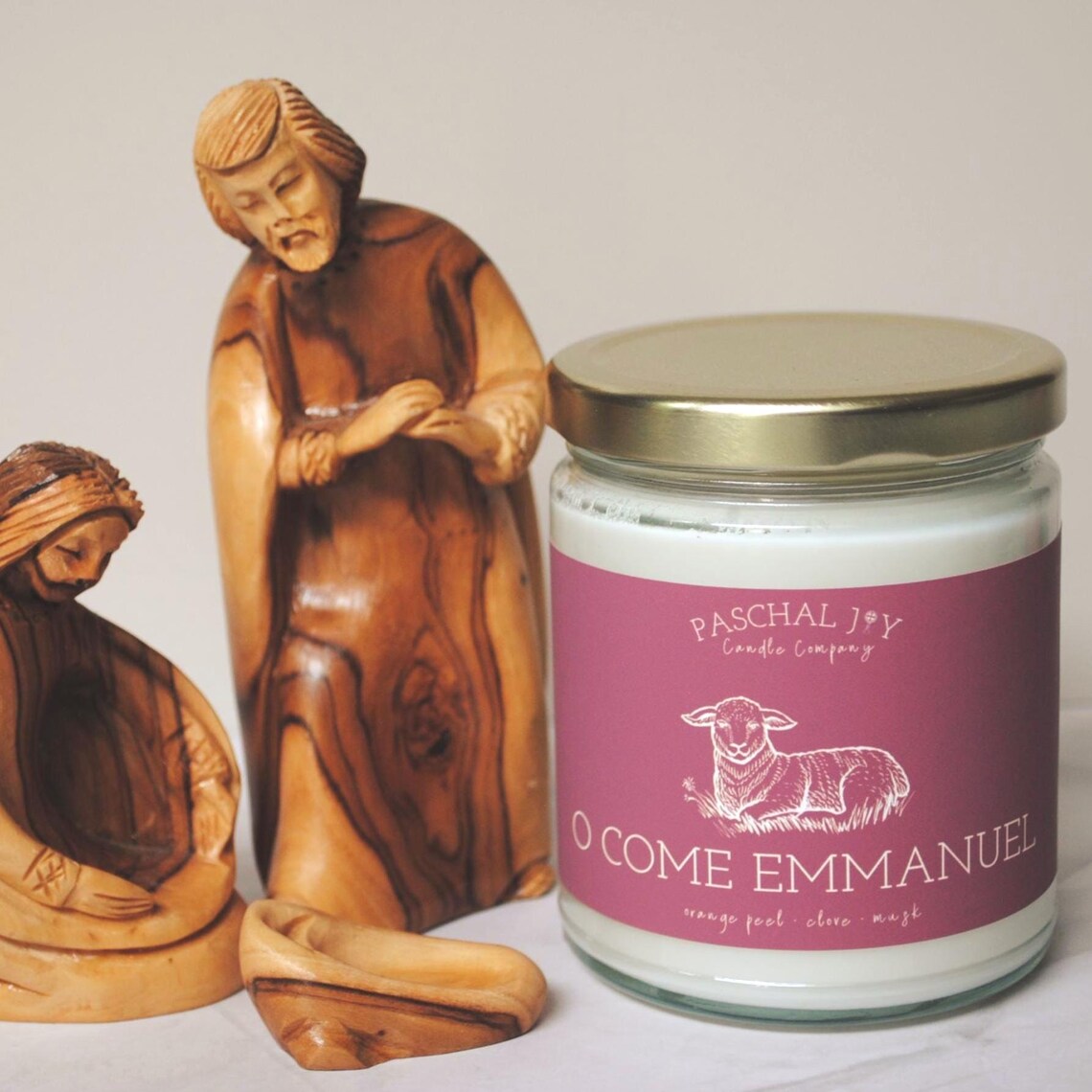 Candle jar with 'O Come Emmanuel' label next to nativity scene figurines on a light background
