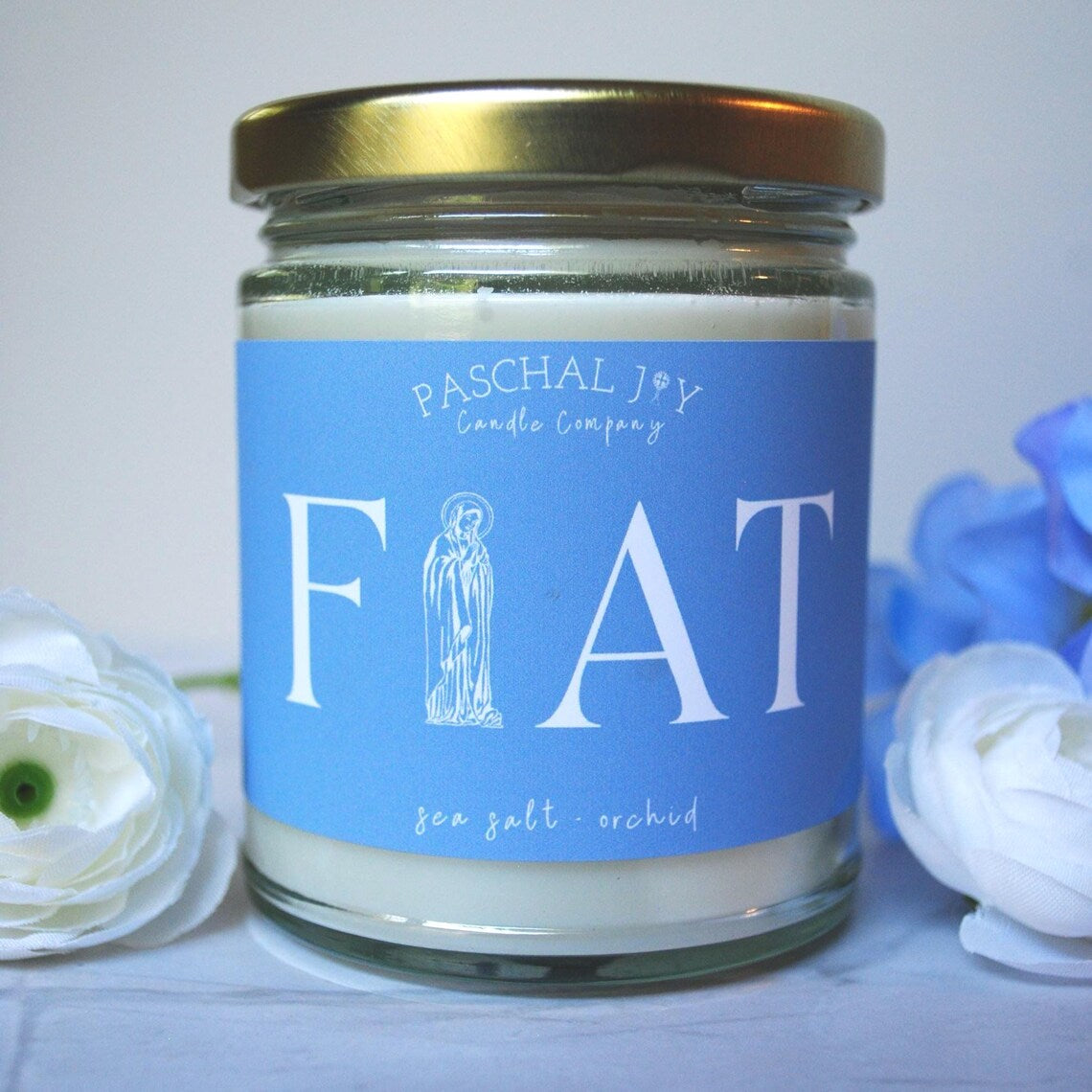 Candle jar with blue label featuring 'Paschal Joy Candle Company' and 'FIAT' text, surrounded by flowers.