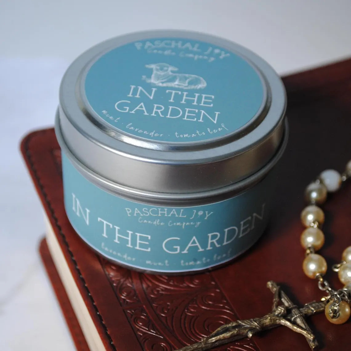 Candle tin labeled 'In the Garden' by Paschal Joy on a textured surface with rosary