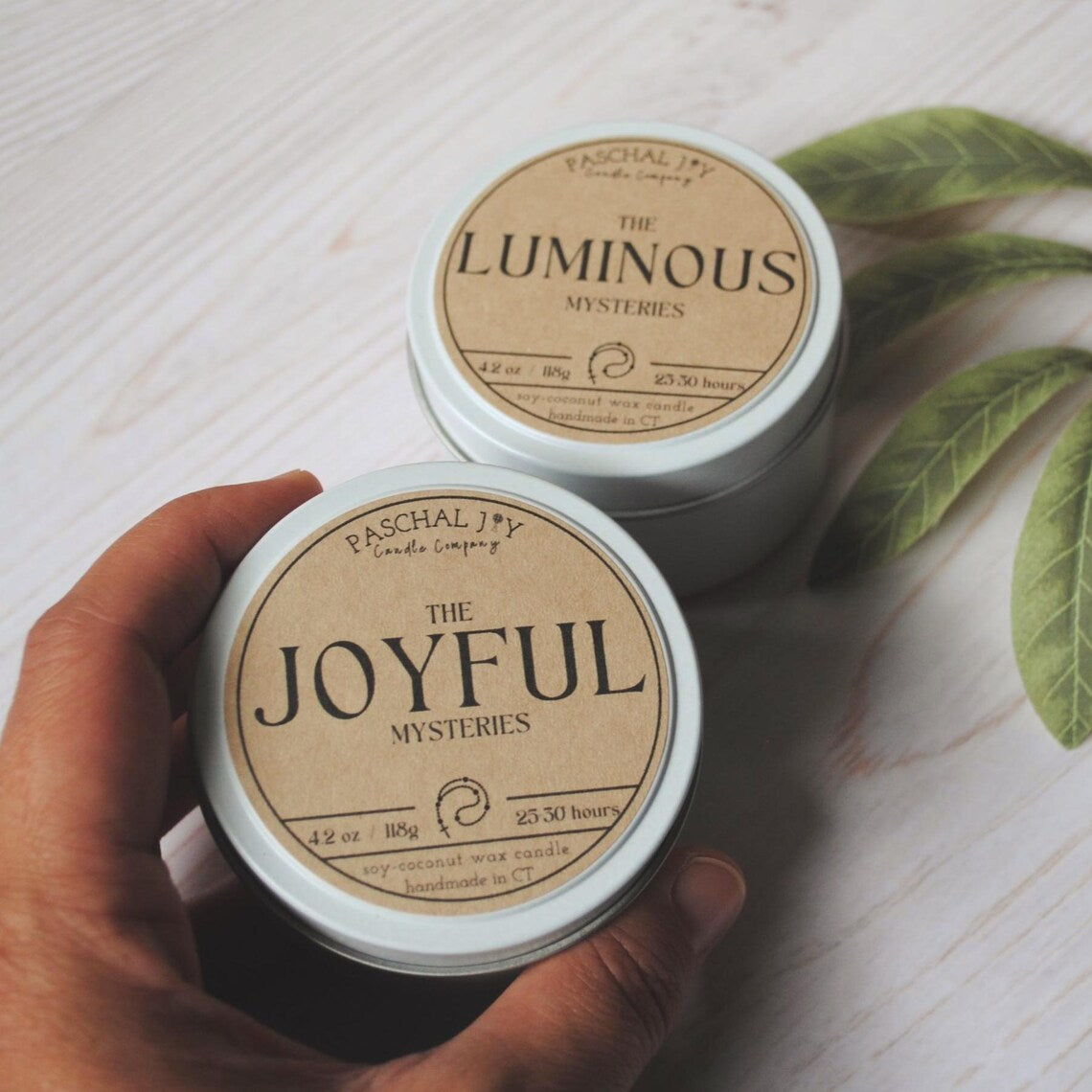 A hand is holding a JOYFUL scented candle tin. The luminous candle tin is in the background