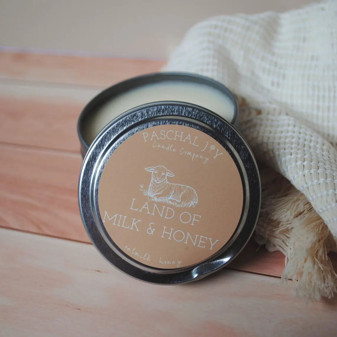 Land of Milk and Honey Candle Tin. Soft white cloth in background with a brown wood ground.