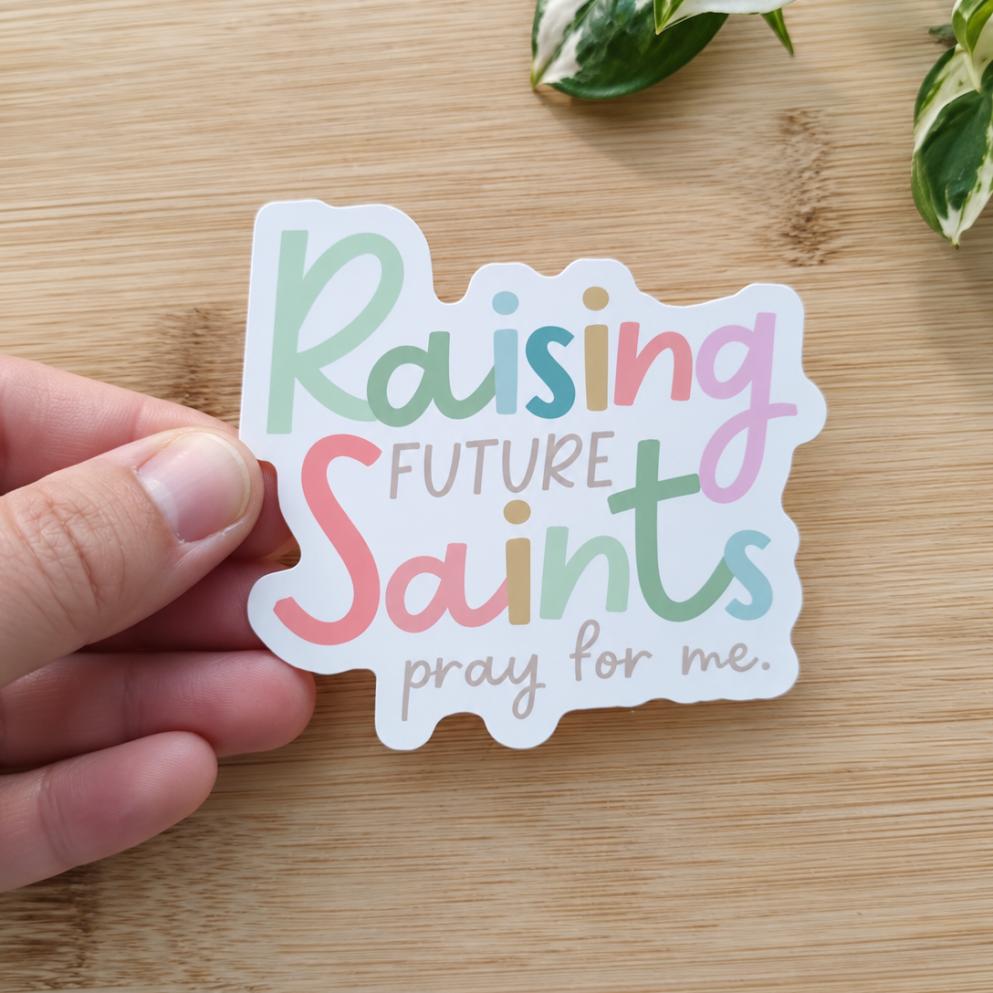 A hand holds a die-cut sticker that reads 'Raising Future Saints pray for me.' in pastel multicolor lettering on a light wood surface with green plant leaves in the corner.