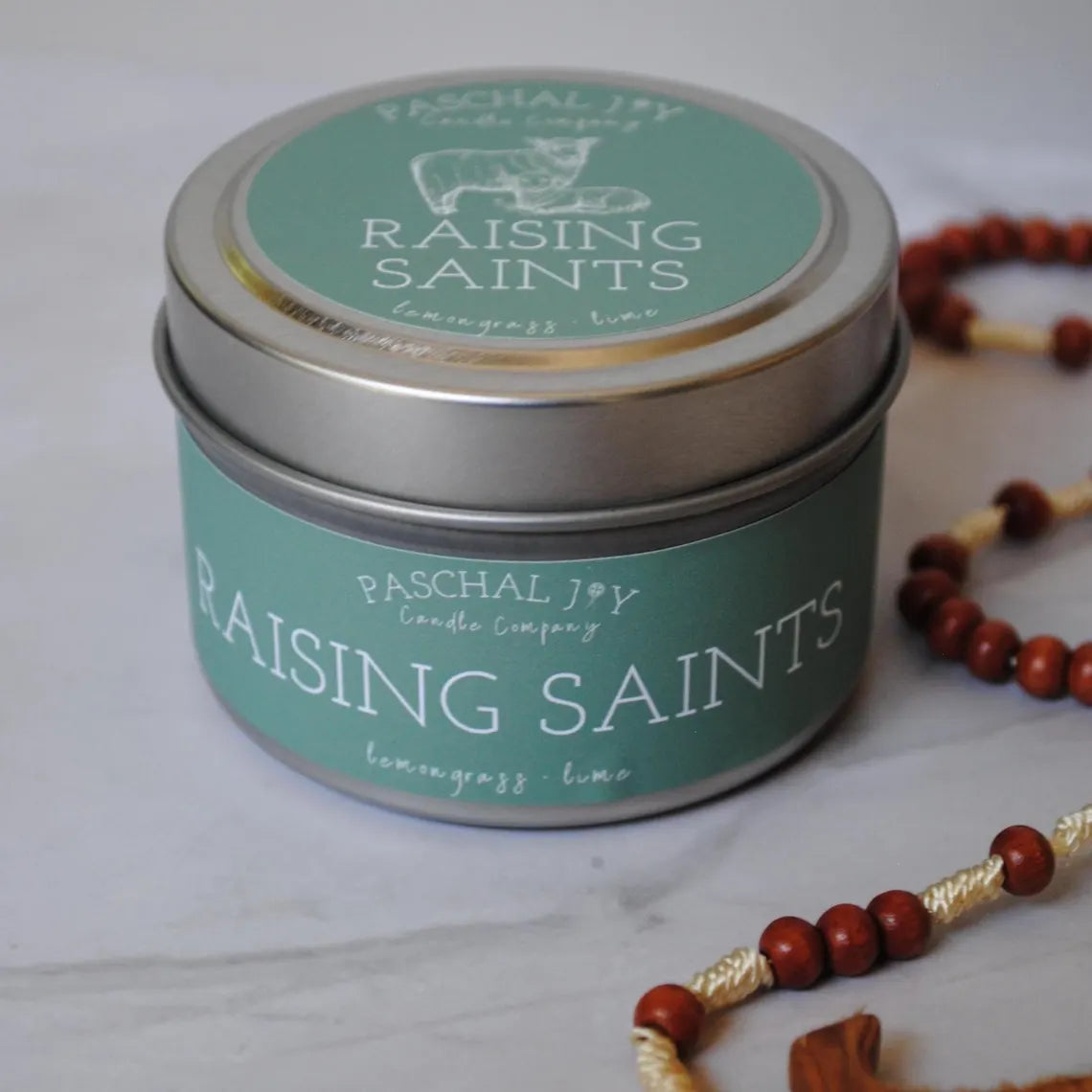 Raising Saints Candle Tin with rosary and grey marble background