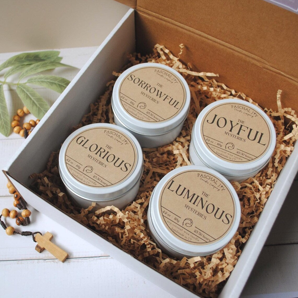 Rosary Candle Collection - white tins with a rosary on a grey background inside of a giftbox with brown crinkle paper
