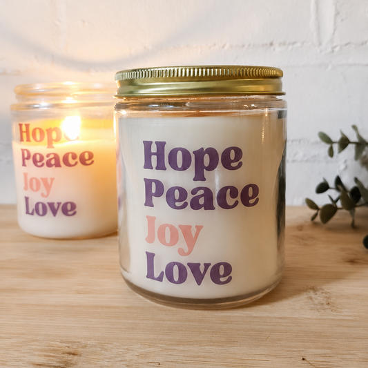 A glass jar candle with a gold metal lid sits on a light wood surface in front of a white brick wall. The candle has a cream-colored wax insert with the words 'Hope Peace Joy Love' printed vertically in purple and pink.
