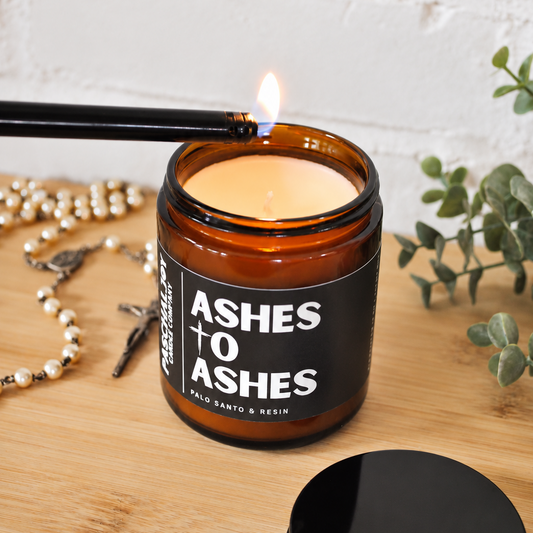 An amber glass jar candle labeled 'ASHES + O ASHES' by Palo Santo & Reed is lit with a black lighter on a wooden surface, with a black lid in front and a white brick wall background.