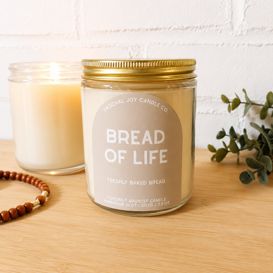 Bread of Life Jar Candle wiht tan label, "freshly baked bread" with rosary beads in background. Wood surface and white brick background.