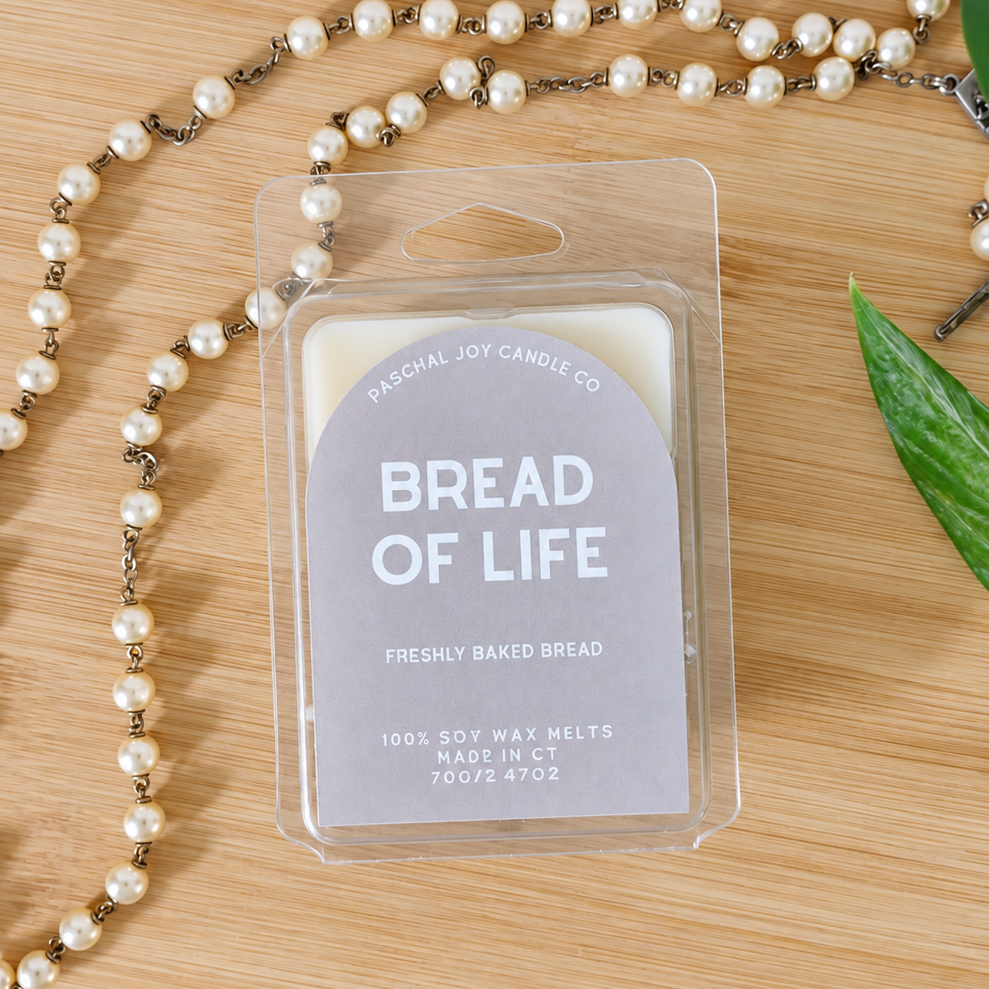A packaged wax melt labeled 'BREAD OF LIFE' by Paschal Joy Candle Co, placed on a light wood surface with a pearl necklace and a green leaf prop.