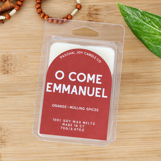 A packaged soy wax melt from Paschal Joy Candle Co. labeled 'O Come Emmanuel' in orange and mulling spices, shown on a light wood surface with decorative beads and a green leaf.