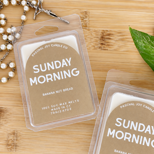 Two clear plastic wax melts hang packs labeled 'Sunday Morning' and 'Banana Nut Bread' by Paschal Joy Candle Co, placed on a light wood surface with a pearl necklace and a green leaf as styled props.