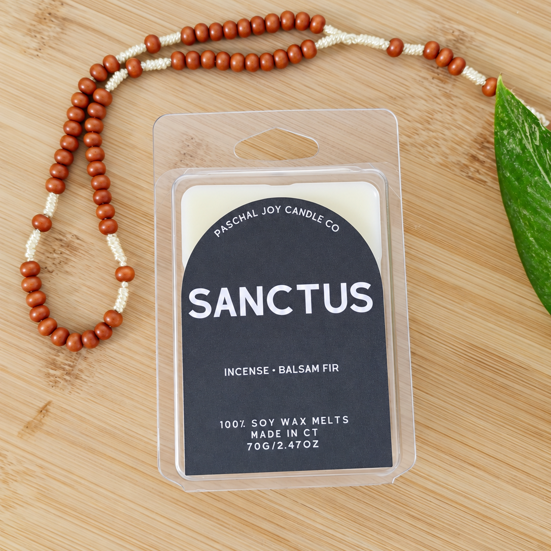 A packaged candle wax melt labeled 'SANCTUS' by Paschal Joy Candle Co., placed on a light wood surface with a beaded necklace and a green leaf nearby.