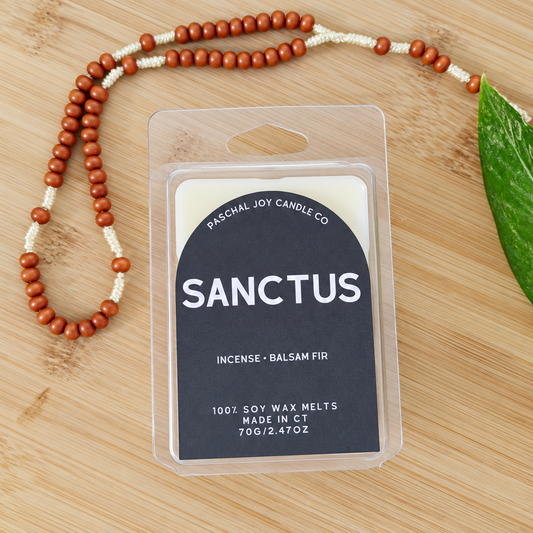 A packaged candle wax melt labeled 'SANCTUS' by Paschal Joy Candle Co., placed on a light wood surface with a beaded necklace and a green leaf nearby.