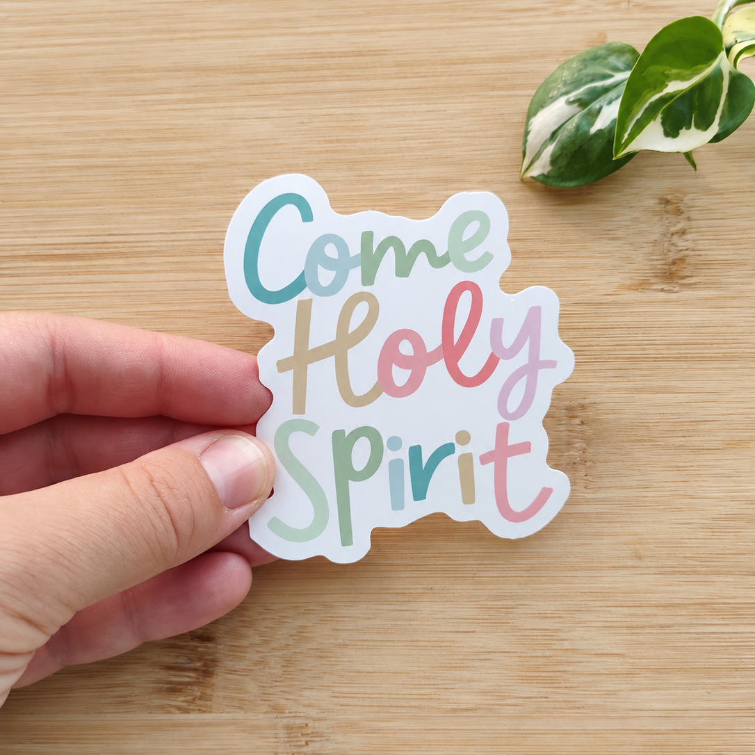 A hand holds a die-cut sticker that reads 'Come Holy Spirit' in pastel blue, peach, and pink lettering on a white background, placed on a light wood surface with a small green plant leaf in the corner.
