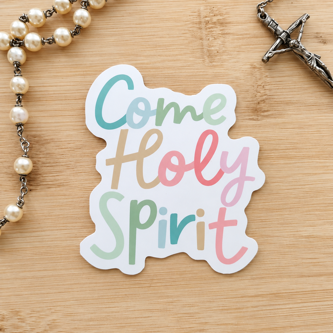 A die-cut sticker with the words 'Come Holy Spirit' in pastel-colored script on a white background, placed on a light wood surface next to a pearl rosary with a silver crucifix.