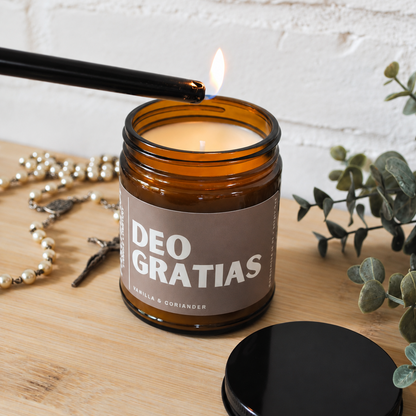 An amber glass jar candle labeled 'DEO GRATIAS' is lit with a black lighter on a wooden surface, with a black lid placed beside it and decorative beads and greenery in the background.