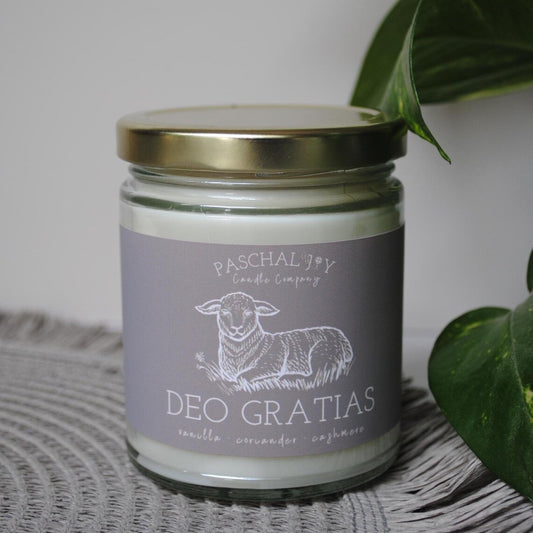 Candle jar with 'Deo Gratias' label and sheep illustration on a textured surface with plant leaves.