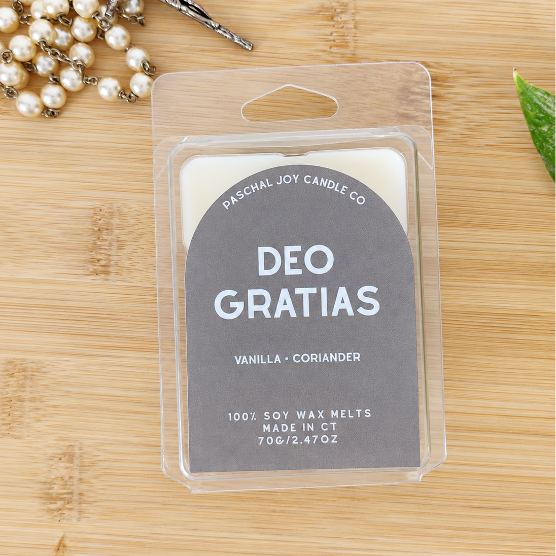 Deo Gratias wax melts in rown arched packaging. On a wooden background with a pearl rosary nearby.