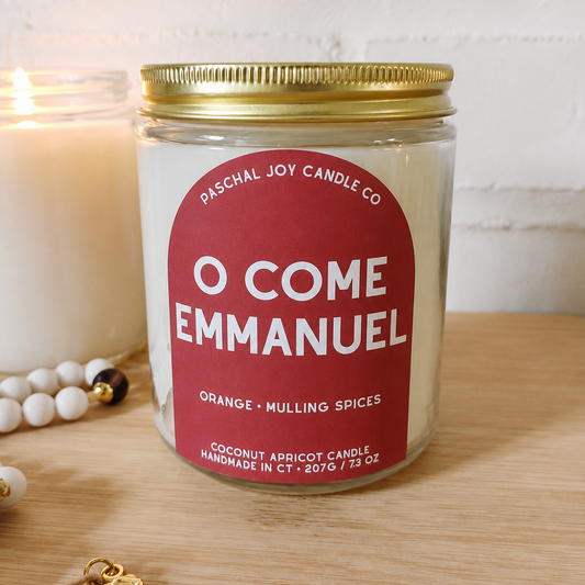 A glass jar candle with a gold metal lid and a red label reading 'O COME EMMANUEL' by Paschal Joy Candle Co, placed on a wooden surface with a blurred white candle and beads in the background.