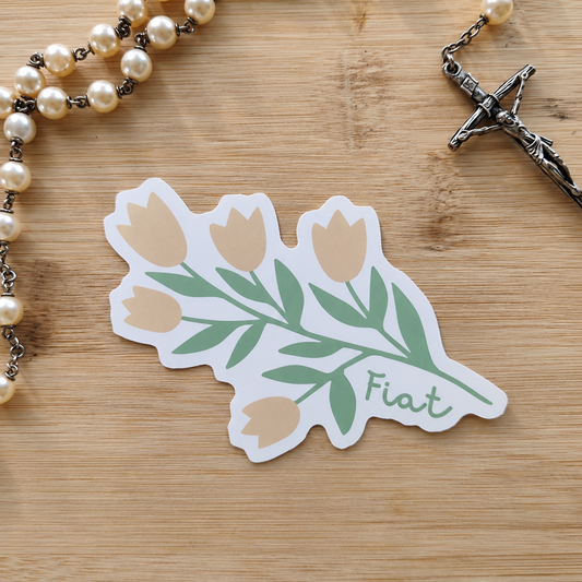A die-cut sticker featuring a bouquet of tulips with green leaves and the word 'Fiat' in script, placed on a light wood surface next to a pearl rosary with a silver crucifix.