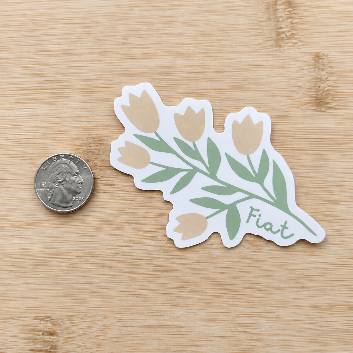 A die-cut sticker featuring tulips with the word 'Fiat' on a light wood surface next to a US quarter for scale.