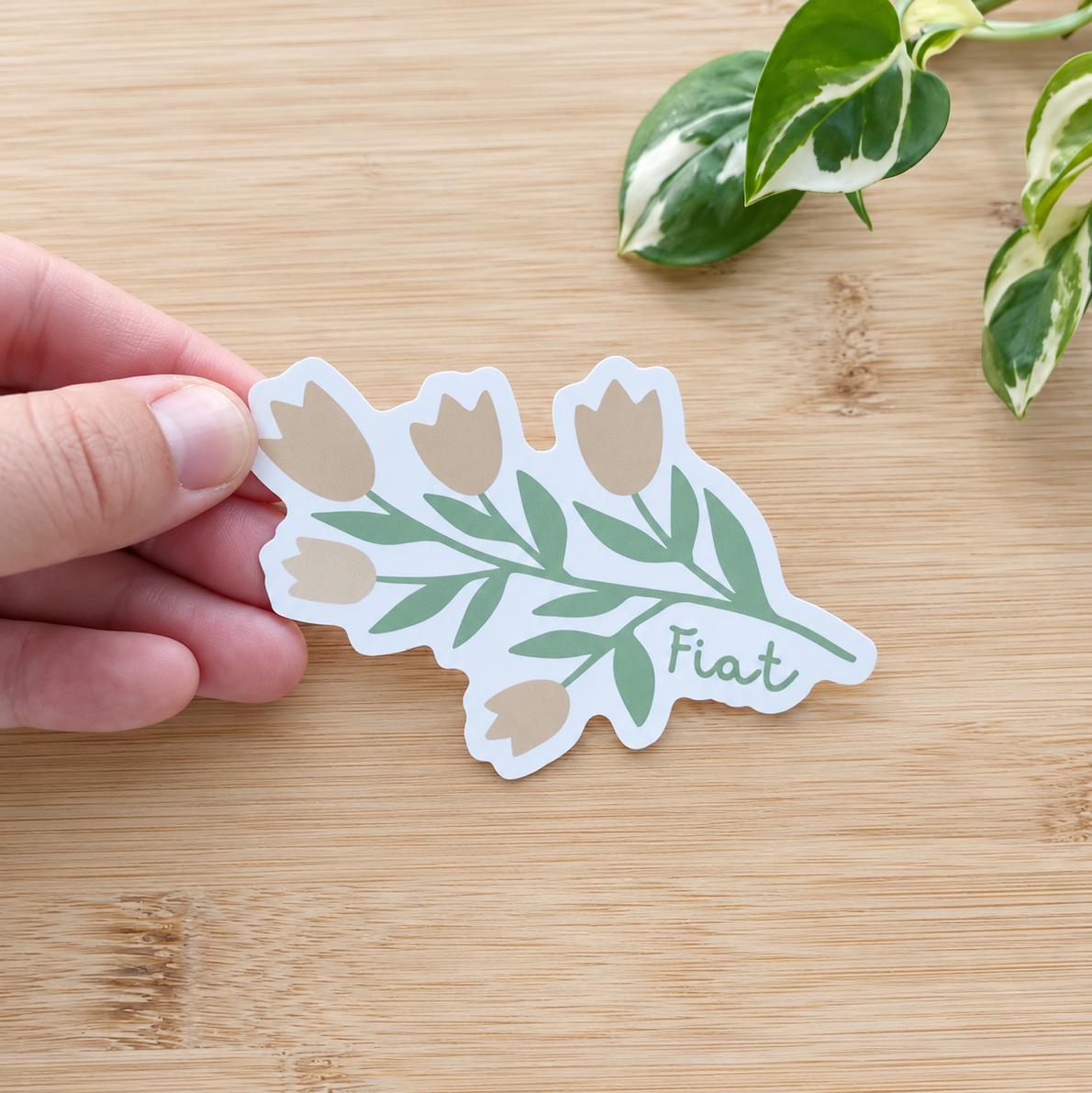 A hand holds a die-cut sticker featuring a botanical design with tulips, green leaves, and the word 'Fiat' on a light wood surface with a green plant in the background.