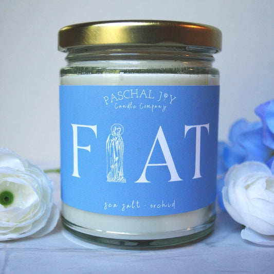 Candle jar with blue label featuring 'Paschal Joy Candle Company' and 'FIAT' text, surrounded by flowers.