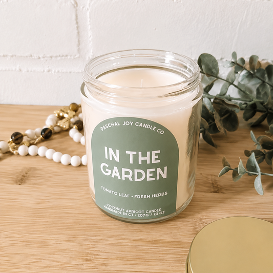 A glass jar candle labeled 'IN THE GARDEN' by Fractal Joy Candle Co., placed on a wooden surface with a gold lid beside it, white beads, greenery, and a white brick wall in the background.