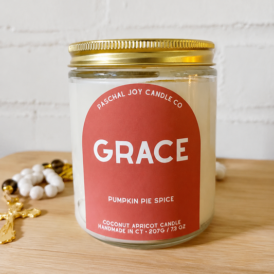 A glass jar candle with a gold metal lid and a red label reading 'GRACE' and 'Pumpkin Pie Spice' by Paschal Joy Candle Co, set on a wooden surface against a white brick wall.