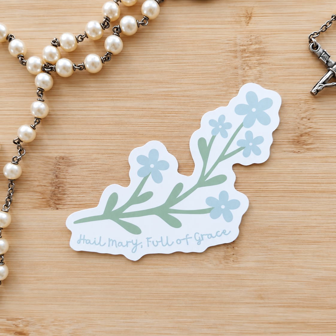A die-cut sticker featuring a light blue and sage green floral branch design with the text 'Hail Mary, Full of Grace' on a white background, styled on a light wood surface next to a pearl rosary.