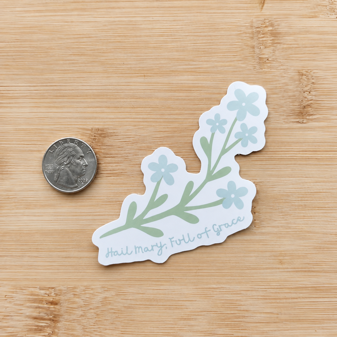 A die-cut sticker featuring a green floral branch with light blue flowers and the cursive text 'Hail Mary, Full of Grace' on a white background, shown on a light wood surface next to a US quarter for scale.