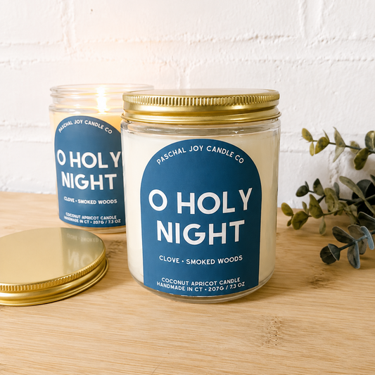 Two glass jar candles with gold lids on a light wood surface against a white brick wall. The front candle has a blue label reading 'O HOLY NIGHT' and 'Pascual Joy Candle Co.