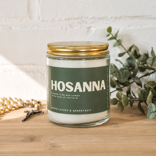 A glass jar candle with a gold metal lid and a dark green label reading 'HOSANNA' sits on a wooden surface against a white brick wall. A gold rosary and green eucalyptus branches are styled around it.