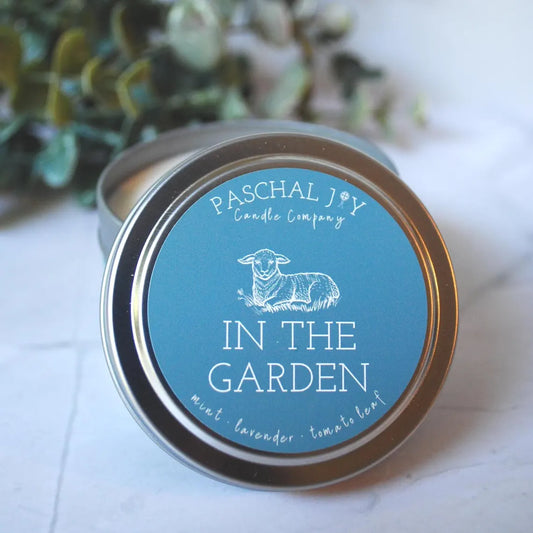 Candle tin labeled 'In the Garden' by Paschal Joy Candle Company on a light surface with greenery in the background.