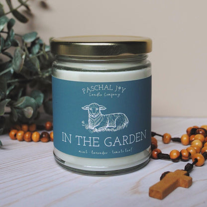 Candle jar labeled 'In the Garden' by Paschal Joy Candle Company with a plant and rosary in the background.