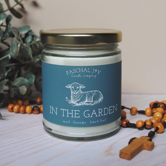 Candle jar labeled 'In the Garden' by Paschal Joy Candle Company with a plant and rosary in the background.