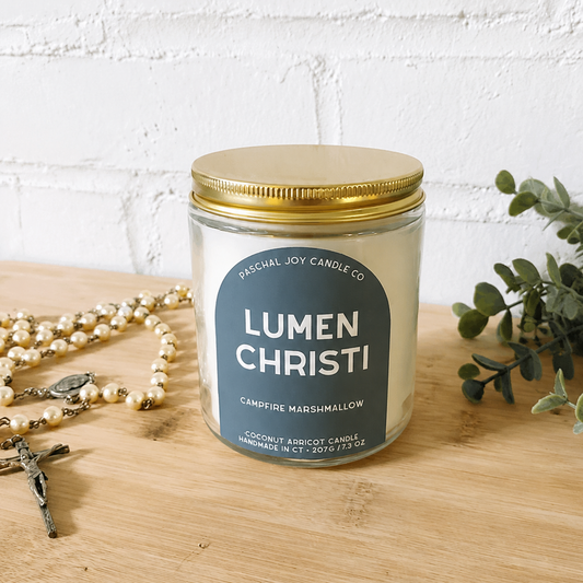 A jar candle labeled 'LUMEN CHRISTI' by Special Joy Candle Co sits on a light wood surface against a white brick wall. The candle has a gold metal lid and a dark blue label.