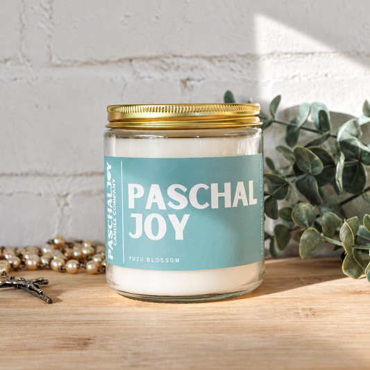A glass jar candle with a gold metal lid and a light blue label reading 'PASCHAL JOY' and 'TRUE BLOOM', placed on a wooden surface against a white brick wall with decorative greenery and beads nearby.