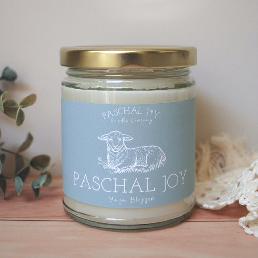 Candle jar with 'Paschal Joy' label featuring a sheep illustration on a wooden surface.