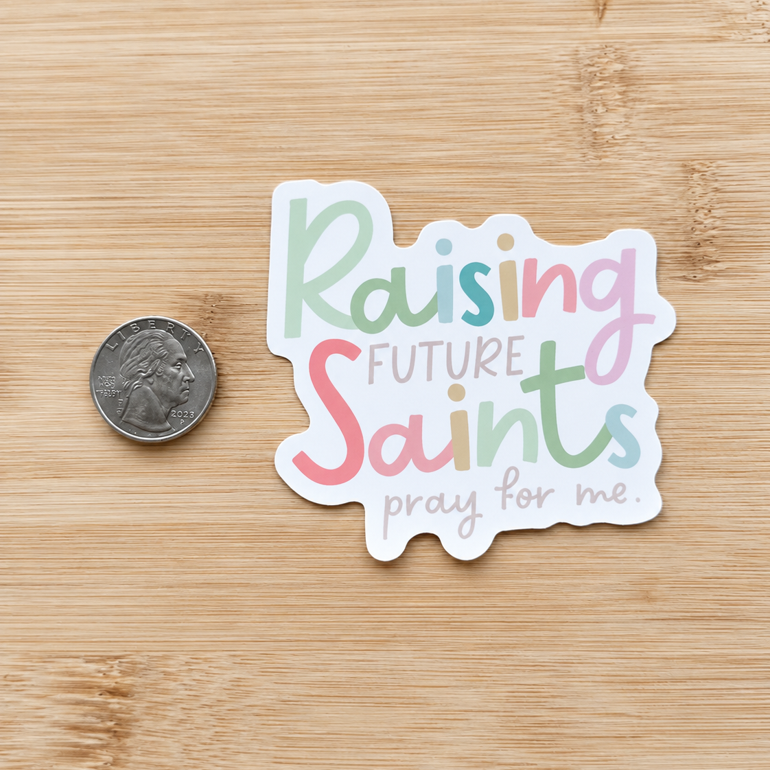 A die-cut sticker with the phrase 'Raising Future Saints pray for me.' in pastel lettering, shown on a light wood surface next to a US quarter for scale.