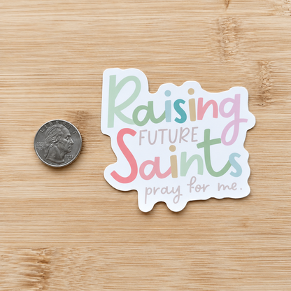 A die-cut sticker with the phrase 'Raising Future Saints pray for me.' in pastel lettering, shown on a light wood surface next to a US quarter for scale.