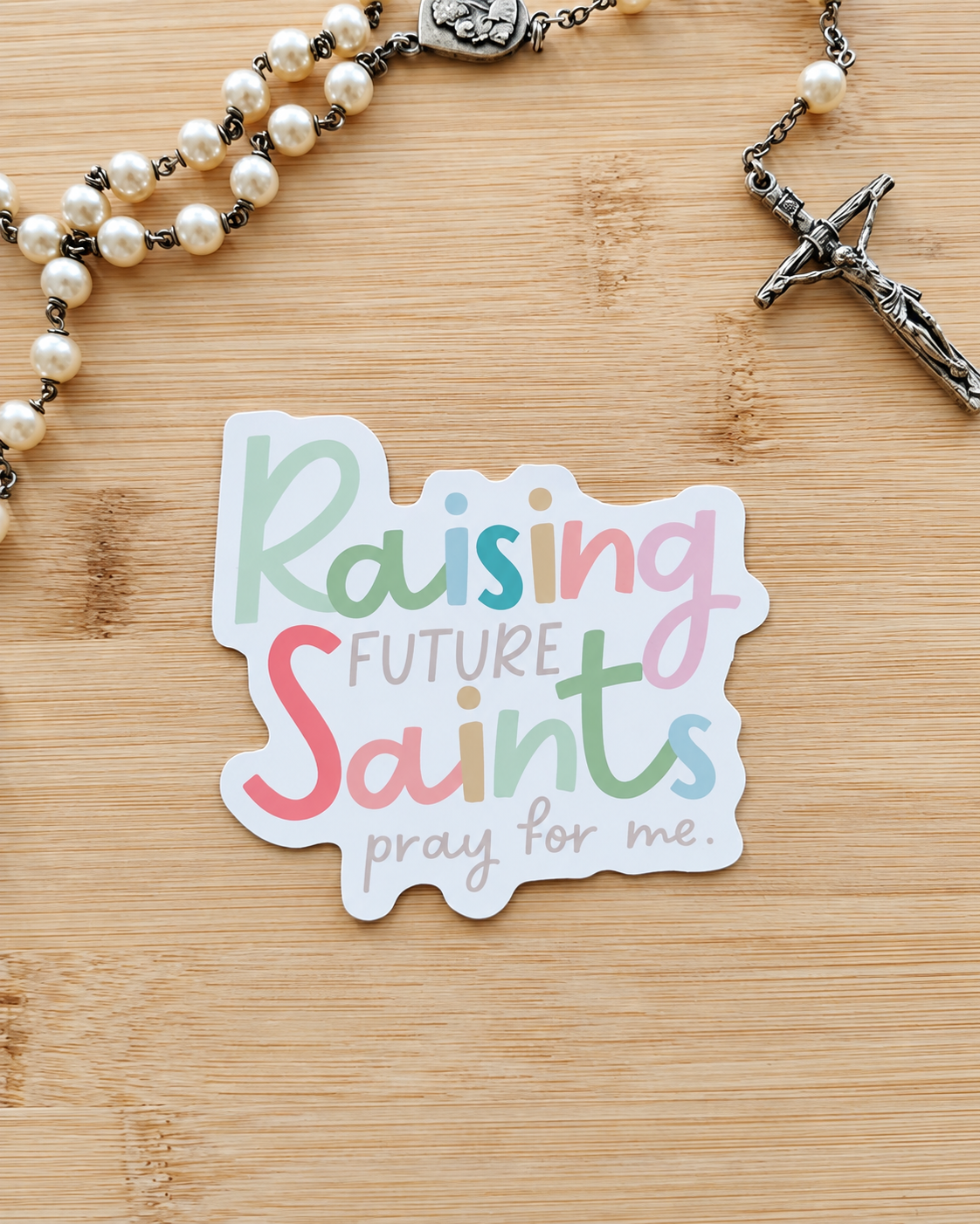 A die-cut sticker with the text 'Raising Future Saints pray for me.' in pastel multicolor lettering, placed on a light wood surface next to a pearl rosary with a silver crucifix.