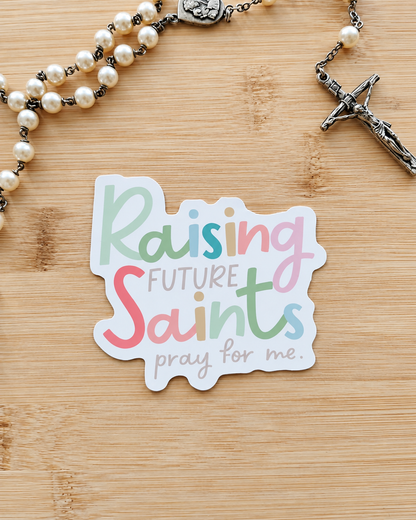 A die-cut sticker with the text 'Raising Future Saints pray for me.' in pastel multicolor lettering, placed on a light wood surface next to a pearl rosary with a silver crucifix.