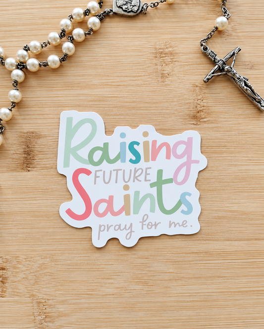 A die-cut sticker with the text 'Raising Future Saints pray for me.' in pastel multicolor lettering, placed on a light wood surface next to a pearl rosary with a silver crucifix.
