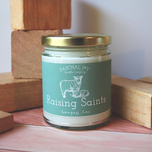 Candle jar labeled 'Raising Saints' by Paschal Joy Candle Company on a wooden surface.