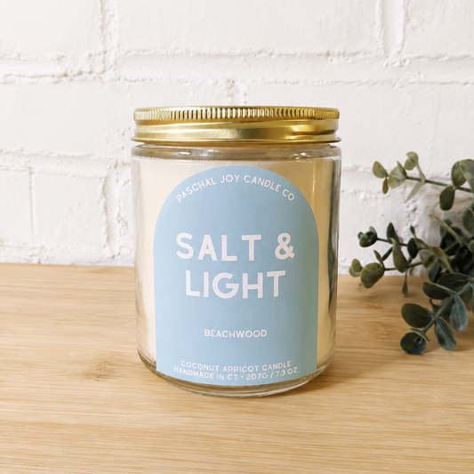 A glass jar candle with a gold metal lid and a light blue label reading 'SALT & LIGHT' and 'Beachwood' by 'Sensual Joy Candle Co', placed on a light wood surface against a white brick wall with greenery on the right.