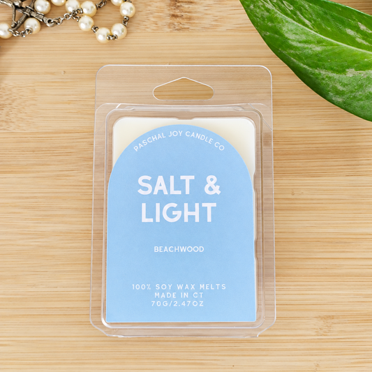 A packaged wax melt product labeled 'SALT & LIGHT' by Paschal Joy Candle Co, placed on a light wood surface with decorative pearl beads and a green leaf around it.