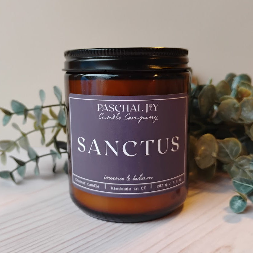 Candle with 'Sanctus' label from Paschal Joy Candle Company on a light surface with greenery.